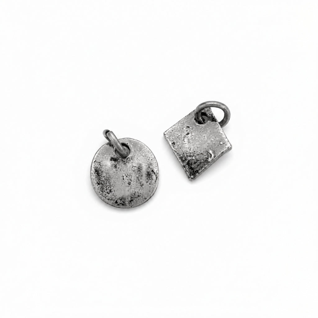 Two textured metal jewelry charms in an antiqued silver finish, one round disc and one square, shown on a white background