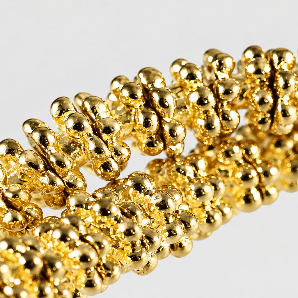 A close-up of a gold-colored beaded chain or bracelet with tightly packed, rounded metallic beads on a plain light gray background.