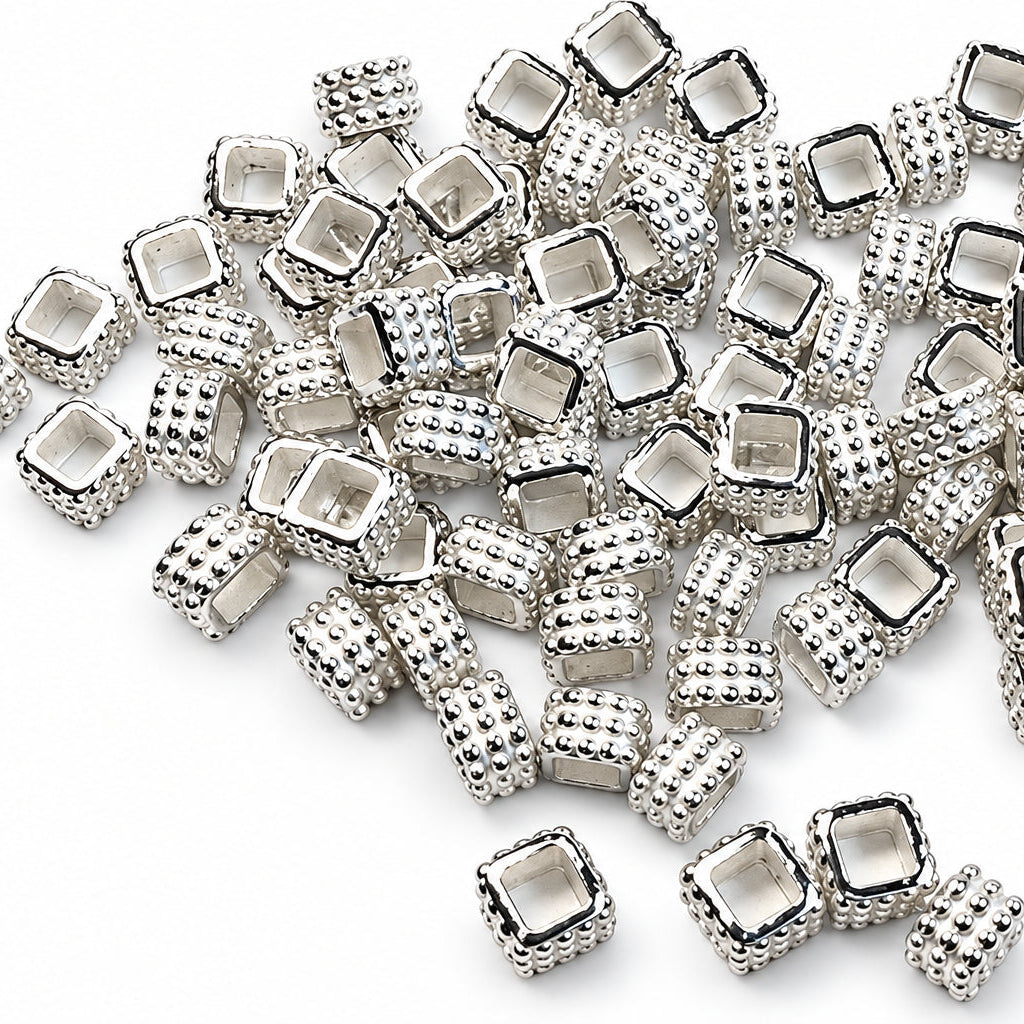 A pile of small silver-toned square spacer beads with textured sides and clear faceted centers, scattered on a plain white background.