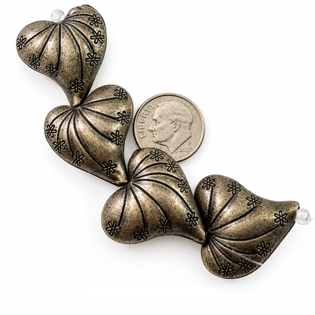 Four metallic heart-shaped beads with a textured bronze finish and floral engravings, shown next to a US dime for scale on a white background.