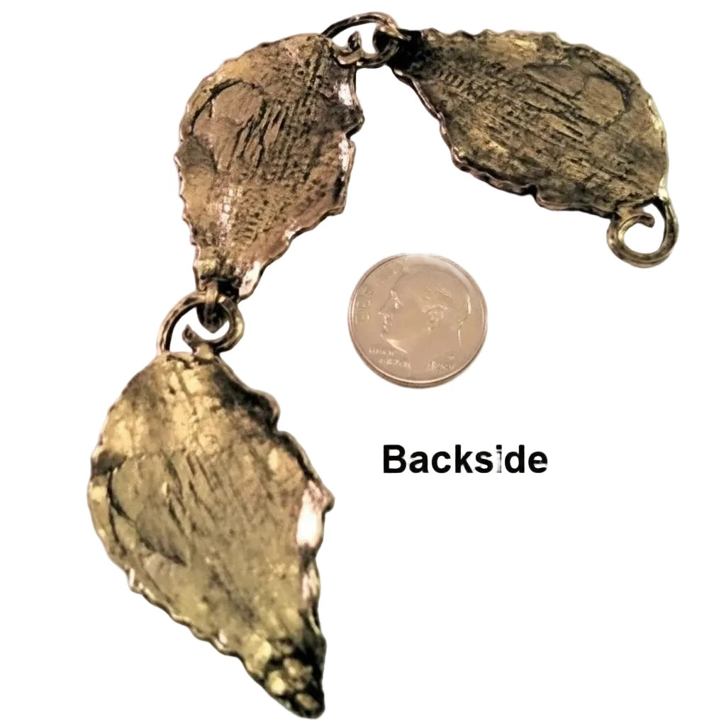 A textured metallic pendant or charm piece shown from the back, with a coin and the text 'Backside' for scale on a plain light background.