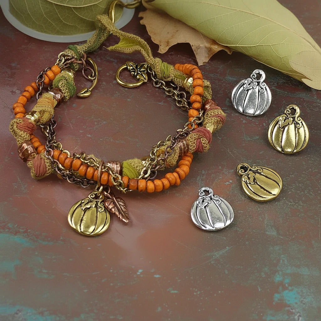 A handmade autumn-themed beaded bracelet with orange beads, green leaves, copper-toned chain links, and pumpkin charms, displayed alongside matching pumpkin charms in silver and gold finishes on a textured brown surface.