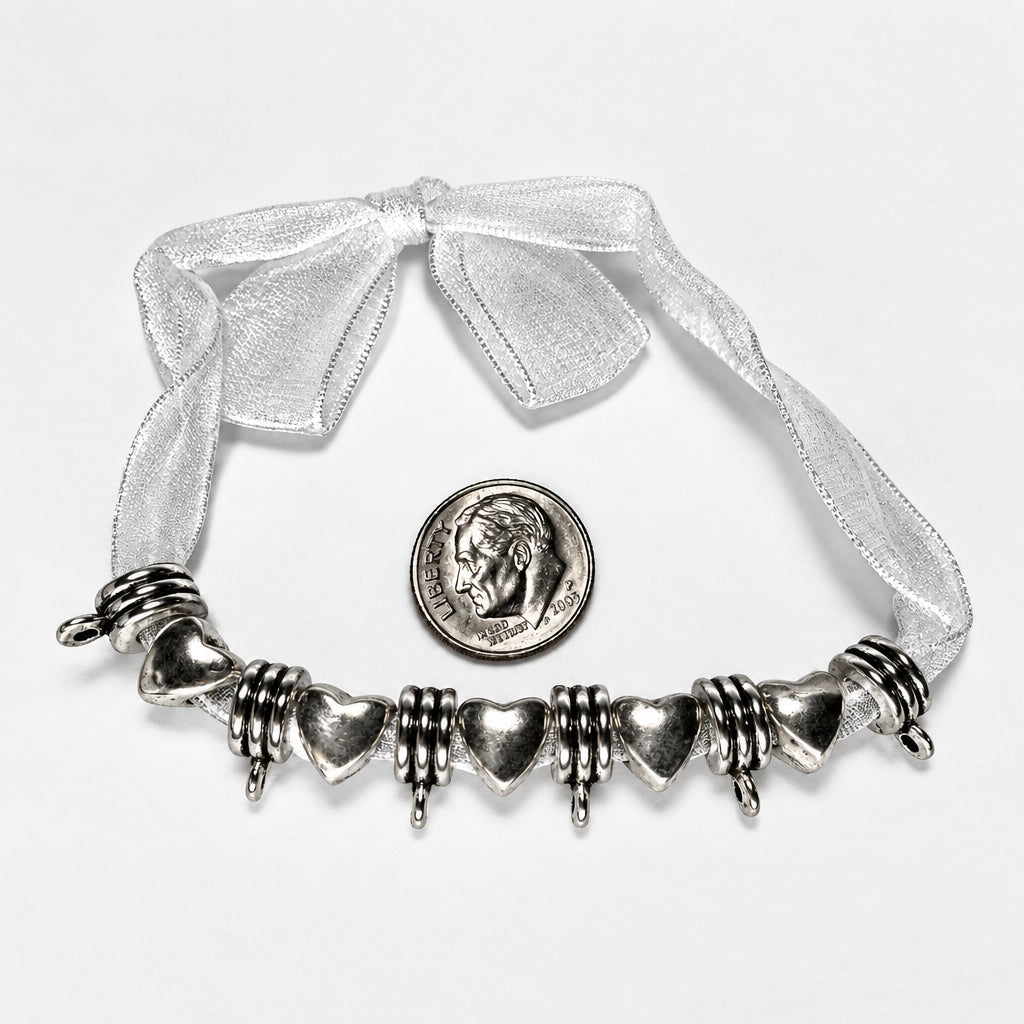 A silver heart charms with textured spiral accents, displayed on a white background with a white sheer ribbon bow and a US quarter coin for scale.
