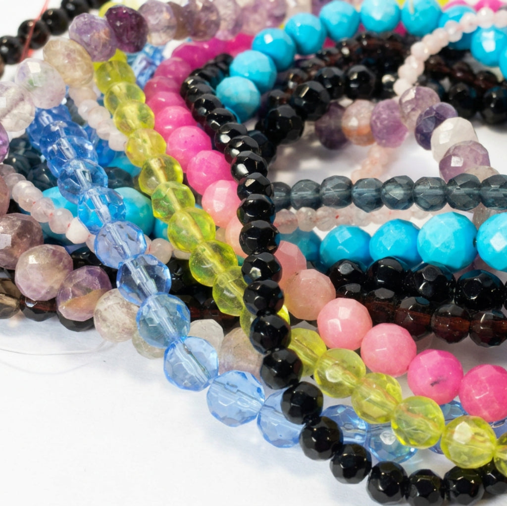 A close-up of multiple strands of faceted round gemstone beads in assorted colors, including turquoise, pink, black, blue, yellow-green, purple, and gray, arranged overlapping on a light surface.