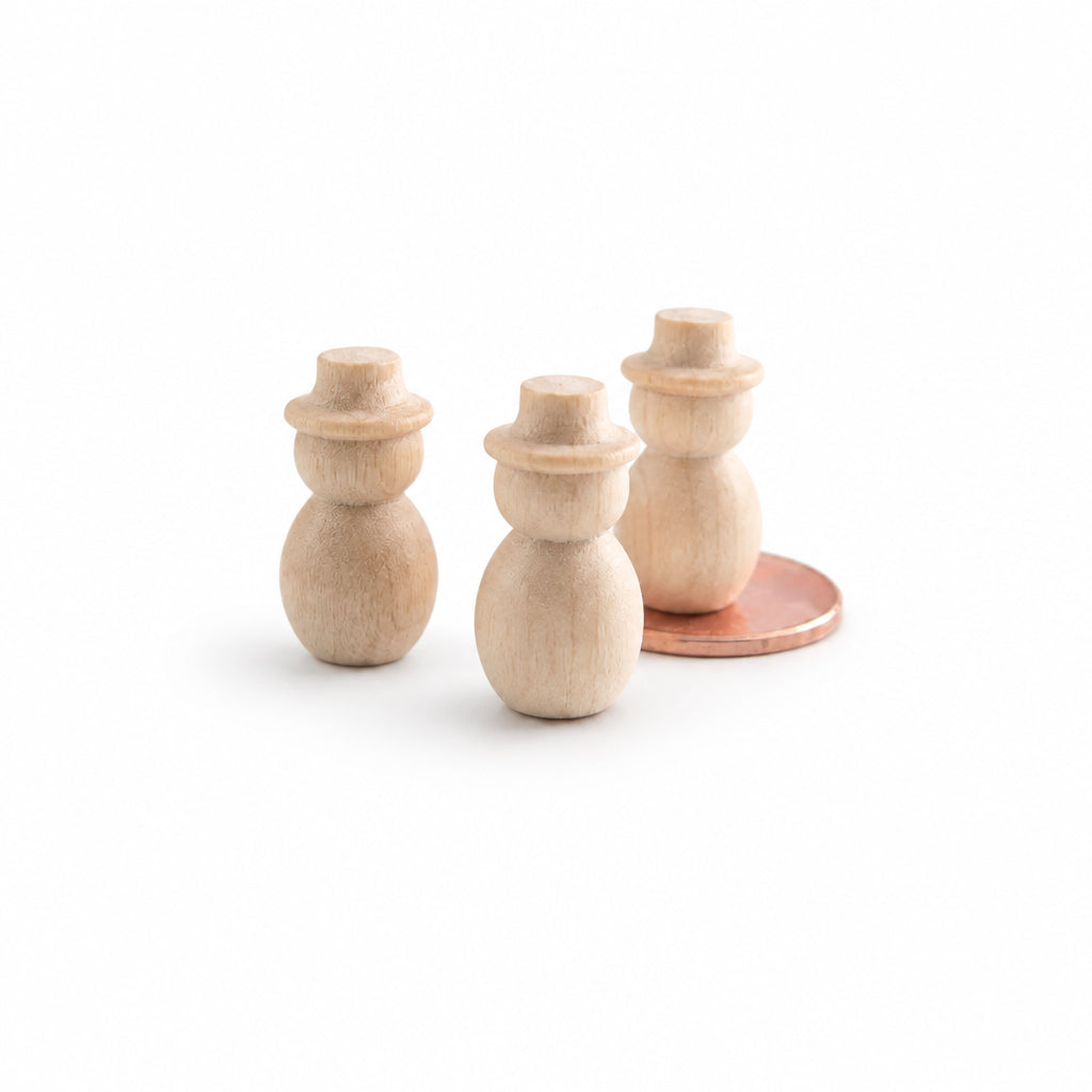 Three small unfinished wooden snowman figurines with a warm natural wood finish, displayed on a clean white reflective surface with a copper coin behind the rightmost figure for scale.