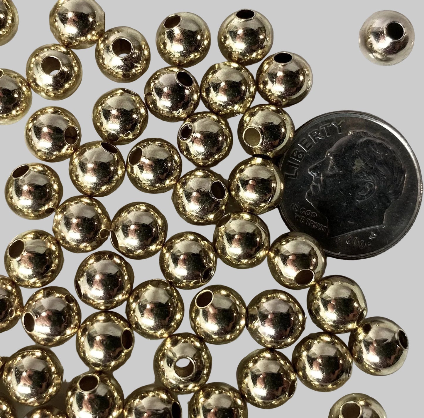 A pile of small metallic gold round beads with central holes, shown on a white surface next to a U.S. dime for scale.
