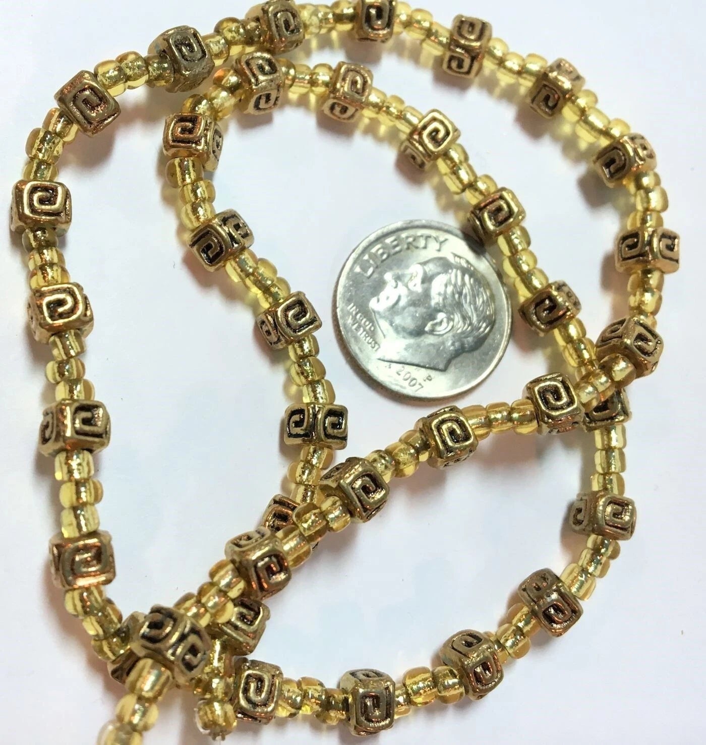 A coiled strand of gold-toned seed beads with metallic square spacers featuring a Greek key pattern, shown next to a silver coin for scale on a plain white background.