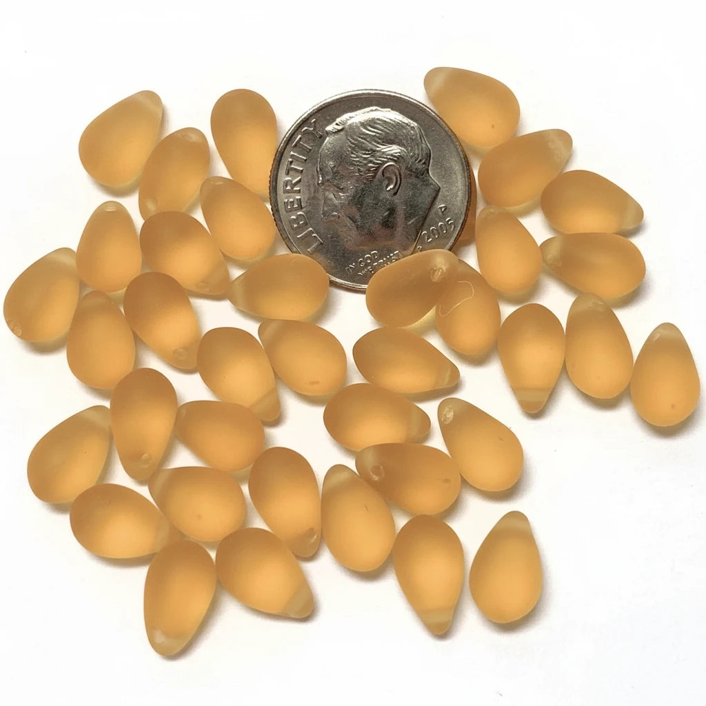 A group of small, teardrop-shaped translucent amber-orange beads scattered on a white background, with a US quarter coin placed near the top for scale.