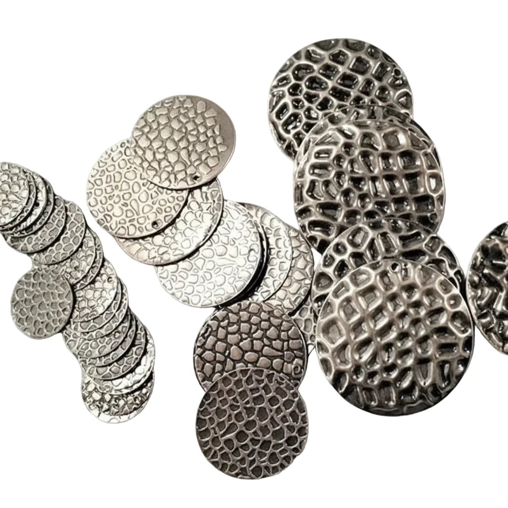 A collection of textured round metal coin-like discs in silver and dark gunmetal finishes, arranged on a light gray textured surface.