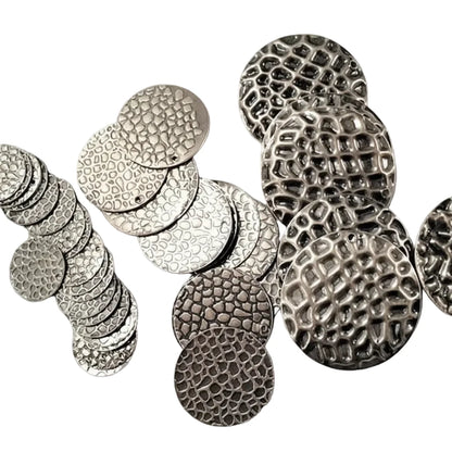 A collection of textured round metal coin-like discs in silver and dark gunmetal finishes, arranged on a light gray textured surface.