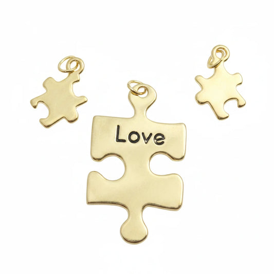 Three gold-toned puzzle piece charms on a light blue textured background, with the center charm engraved with the word 'Love' and a watermark reading 'TheMysticalGardenPath' across the bottom.