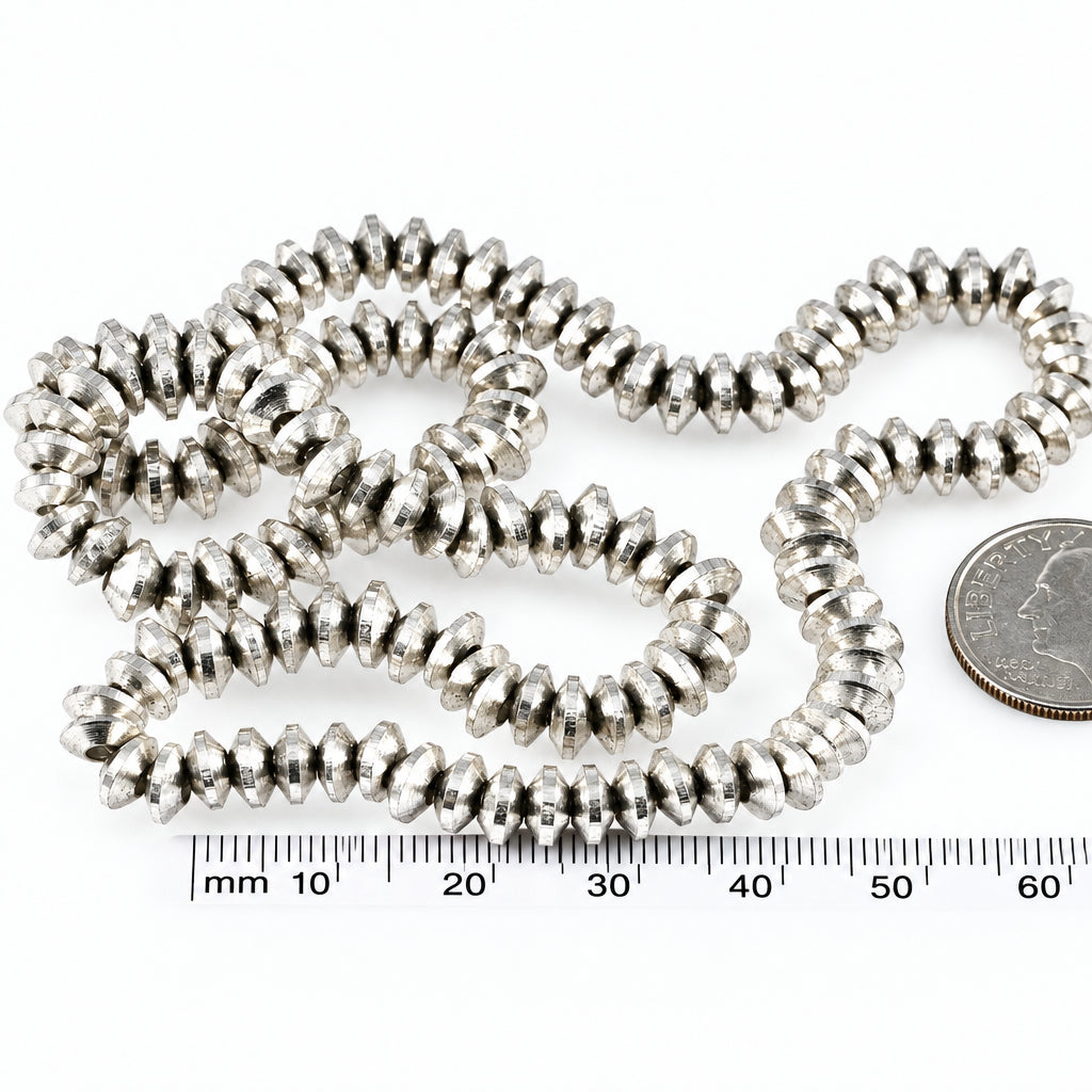 A close-up of a coiled strand of small metallic silver beads or spacers on a white background, with a coin and a millimeter ruler included for scale.