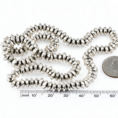 A close-up of a coiled strand of small metallic silver beads or spacers on a white background, with a coin and a millimeter ruler included for scale.
