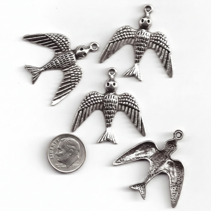 Four silver bird charms with wings spread, shown on a white background with a US quarter coin for scale.