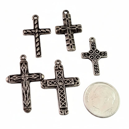 Five silver ornate cross charms with textured details, displayed on a white background next to a US dime for scale.