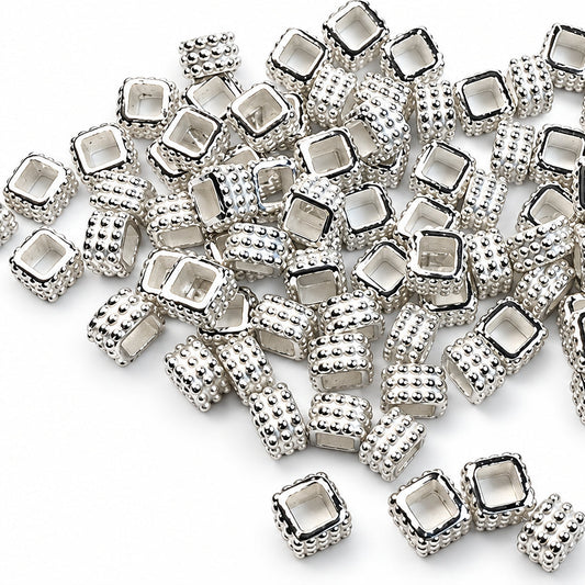 A pile of small silver-toned square spacer beads with textured sides and clear faceted centers, scattered on a plain white background.