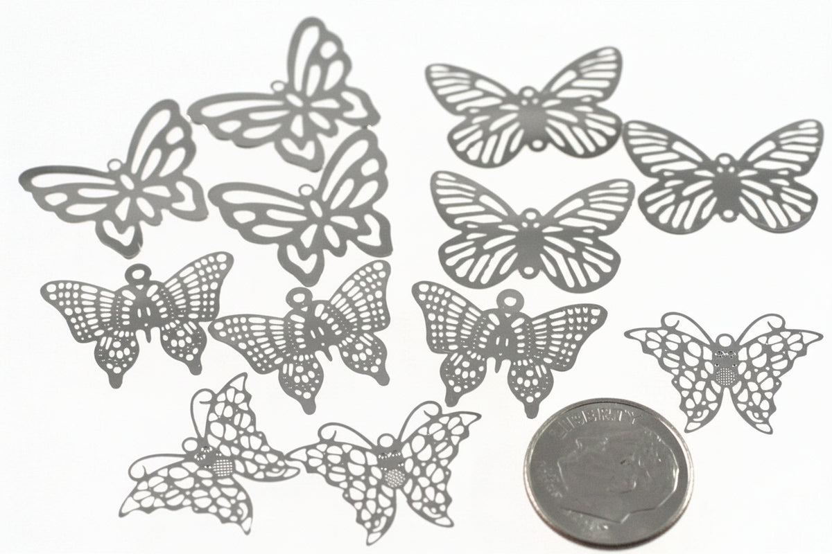 Ten silver butterfly-shaped metal charms or embellishments with intricate lace-like cutouts, arranged on a white background next to a US quarter coin for scale.