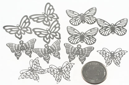 Ten silver butterfly-shaped metal charms or embellishments with intricate lace-like cutouts, arranged on a white background next to a US quarter coin for scale.