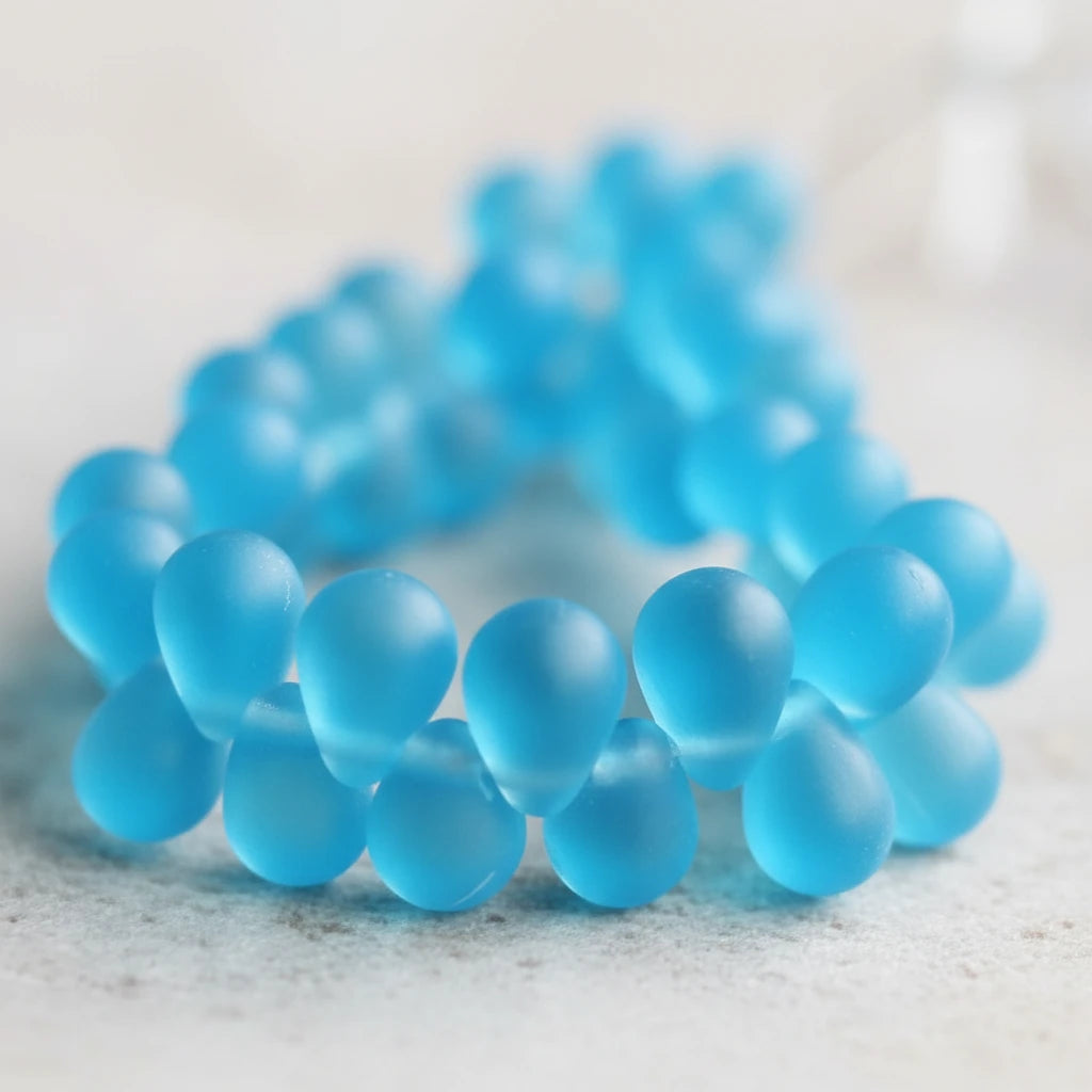 A close-up of a beaded bracelet made of translucent light blue round beads, photographed on a neutral textured surface with a shallow depth of field.