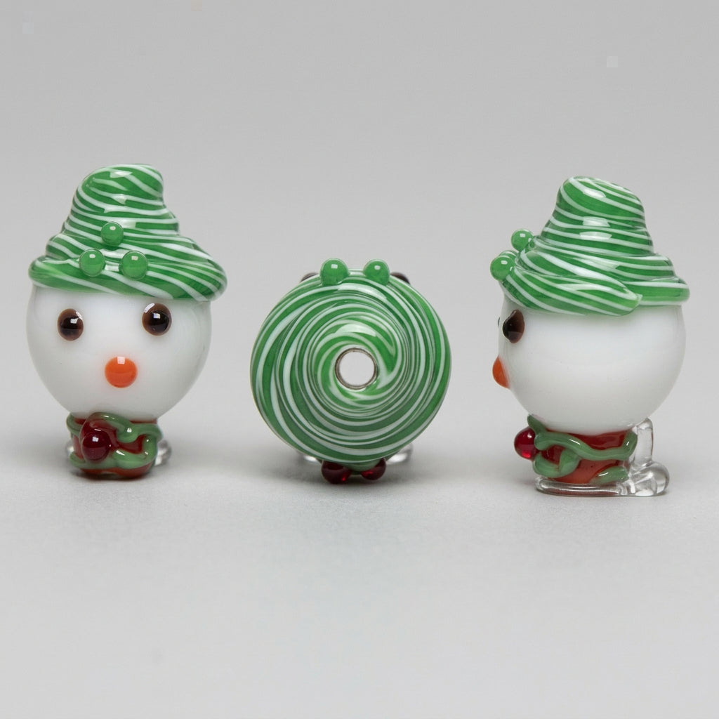 Three views of a festive snowman glass bead on a plain white background: front, back, and side profiles. The bead features a white body, a green and white striped hat, a green swirl body, and a small orange beak.