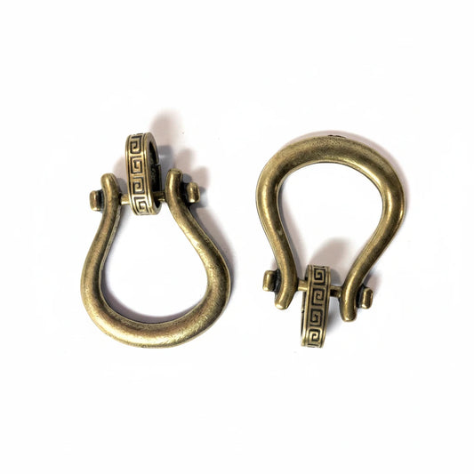 Two antique bronze metal D-rings with Greek key detailing on the clasp, displayed on a white background with a coin for scale on the right.