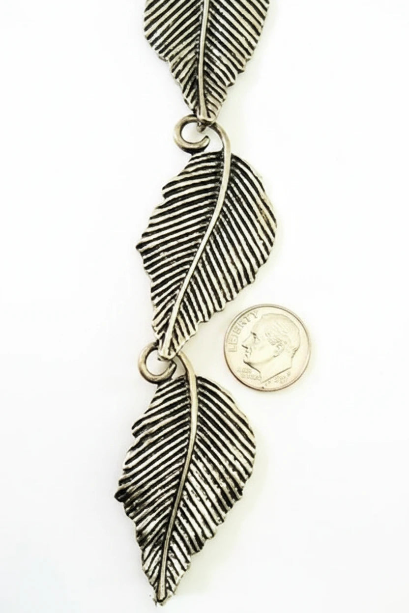 A pair of antique silver feather charms with detailed textured stripes, shown next to a US quarter coin for scale on a white background.