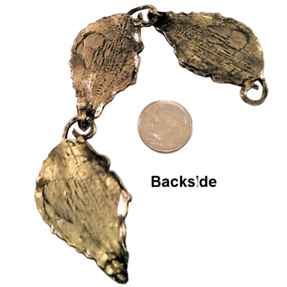 A textured metallic pendant or charm piece shown from the back, with a coin and the text 'Backside' for scale on a plain light background.