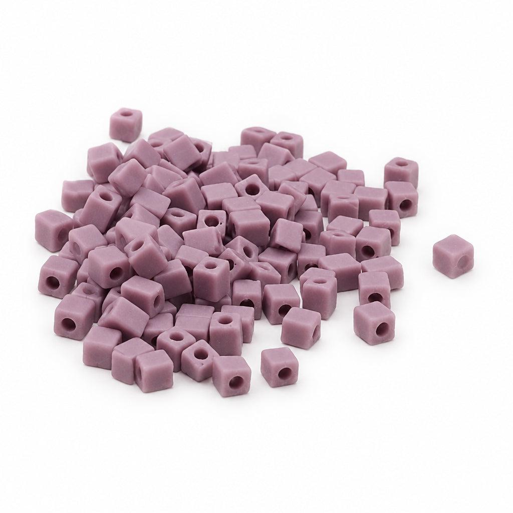 A pile of small lavender-purple cube-shaped seed beads with center holes, scattered on a plain white background.