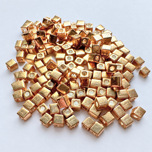 A pile of small metallic gold cube-shaped beads with circular holes, photographed close-up on a light gray background.
