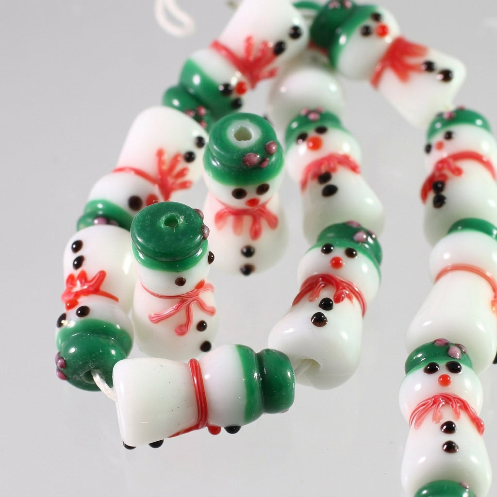 A strand of handmade glass beads shaped like snowmen, featuring white bodies with green hats and boots, red scarves, and black facial details, shown on a white background with a coin for scale.