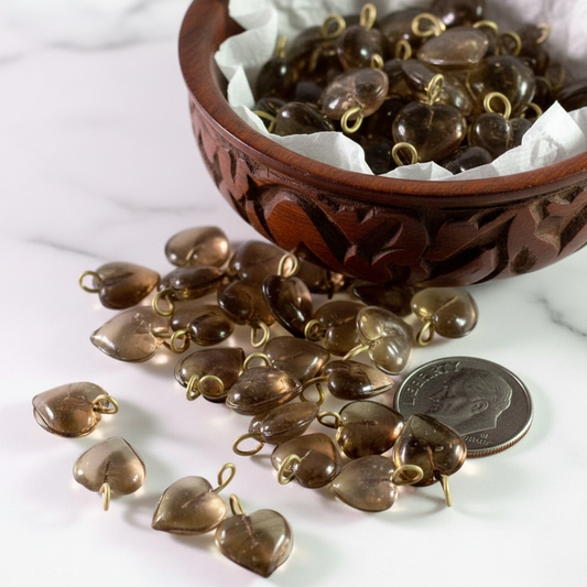 Heart-shaped smoky brown glass beads with gold-tone loops, shown spilling from a carved wooden bowl onto a white marble surface, with a coin for scale.