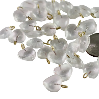 Clear glass heart-shaped beads with small gold metal loops, scattered on a light pink background with a coin for scale.