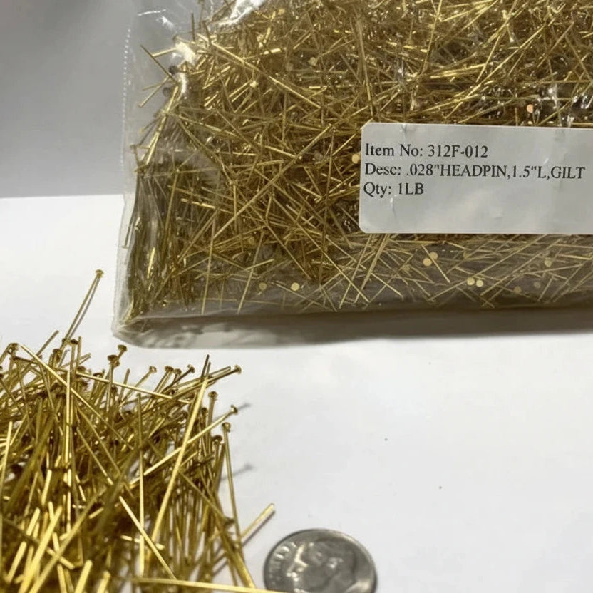 A clear plastic bag filled with gold-headed pins, with a few pins spilled in the foreground and a white label showing product details. A coin is placed near the bottom right for scale.