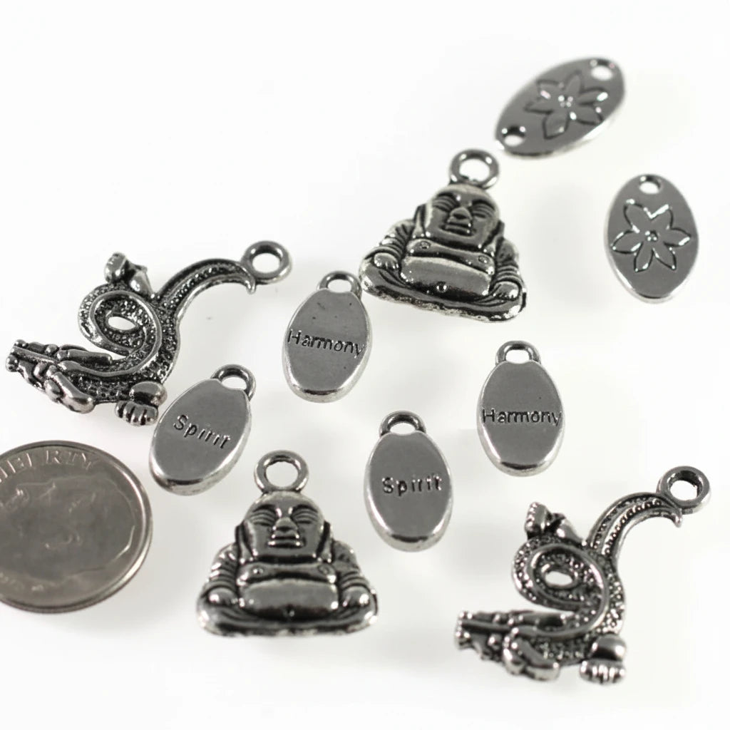 A set of silver-toned metal jewelry charms and oval tags with engraved words like 'Harmon' and 'Spirit,' plus decorative motifs including a seated Buddha figure, a coiled snake, and a flower, shown on a white background with a coin for scale.