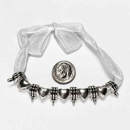 A silver heart charms with textured spiral accents, displayed on a white background with a white sheer ribbon bow and a US quarter coin for scale.