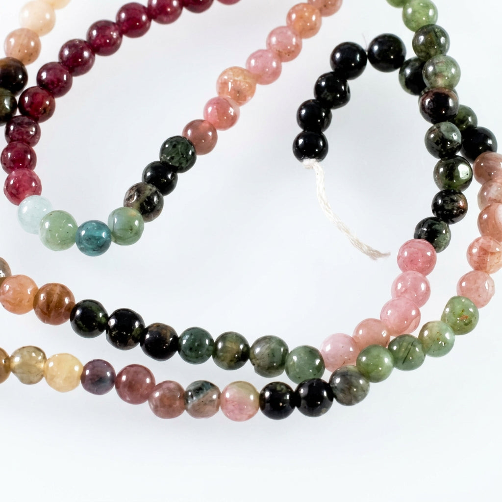 A close-up of a beaded strand made of round polished gemstone beads in multiple colors, including pink, green, black, brown, and pale blue, arranged on a plain light background.