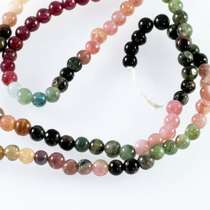 A close-up of a beaded strand made of round polished gemstone beads in multiple colors, including pink, green, black, brown, and pale blue, arranged on a plain light background.