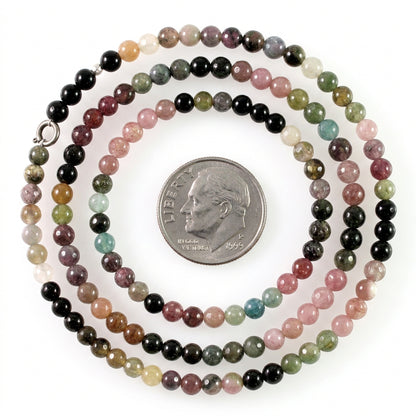 A beaded strand necklace made of small polished round beads in multiple colors including black, pink, green, brown, and white, shown with a silver coin for scale on a white background.