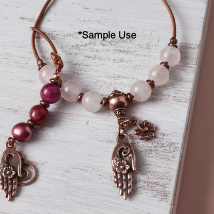 A close-up of a beaded necklace with translucent pale pink beads, a copper-toned central bead, a dangling hand-shaped charm with floral details, and a small flower charm, laid on a distressed white wood surface. Black text at the top reads '*Sample Use'.