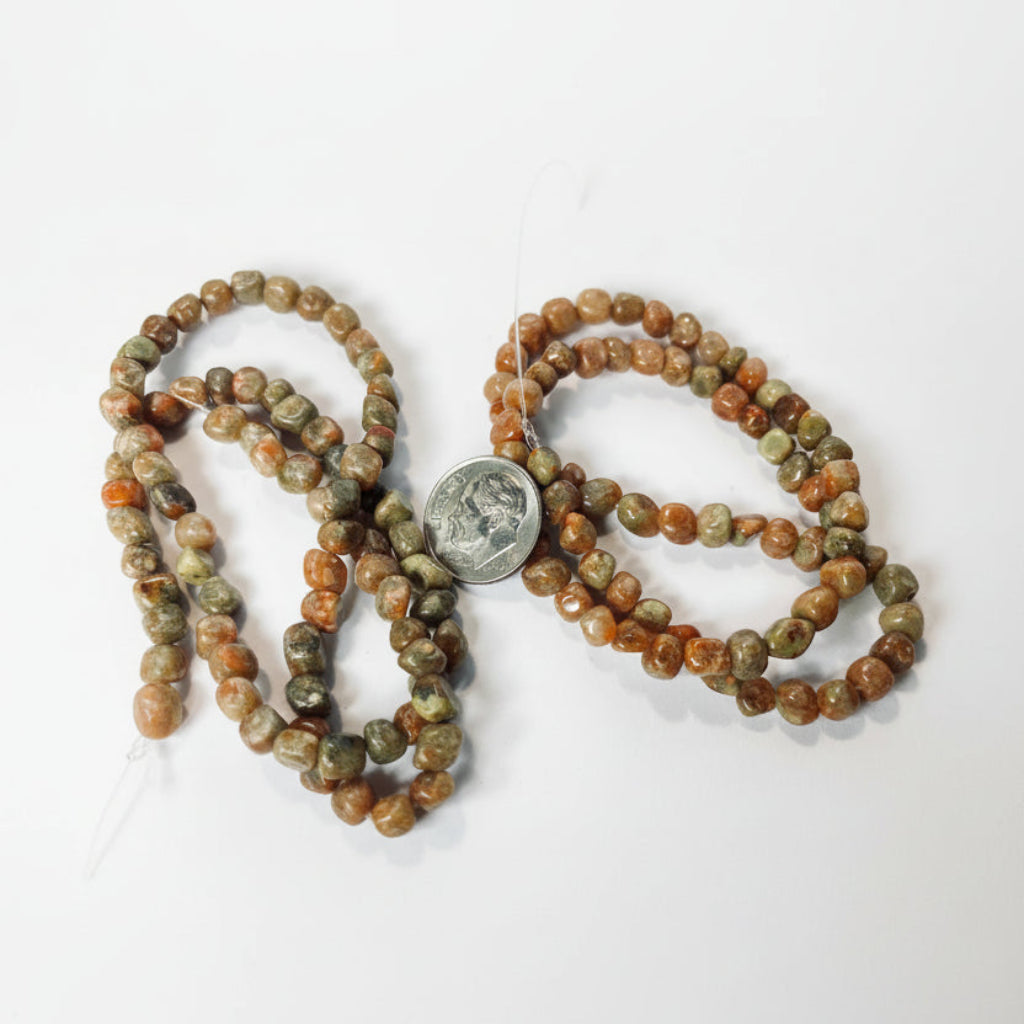 Tumbled stone beads in earthy brown, orange, and green hues, shown draped on a white surface with a US quarter coin in the center for scale.