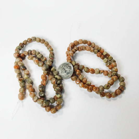 Tumbled stone beads in earthy brown, orange, and green hues, shown draped on a white surface with a US quarter coin in the center for scale.