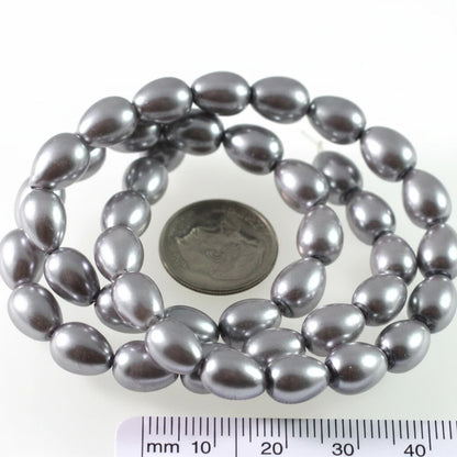 A coiled strand of dark gray teardrop-shaped beads with a metallic sheen, shown next to a US quarter coin for scale and a black measurement ruler at the bottom.
