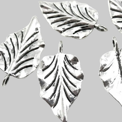 Five silver-toned leaf-shaped jewelry charms with textured leaf veins and hanging loops, arranged on a light gray background.