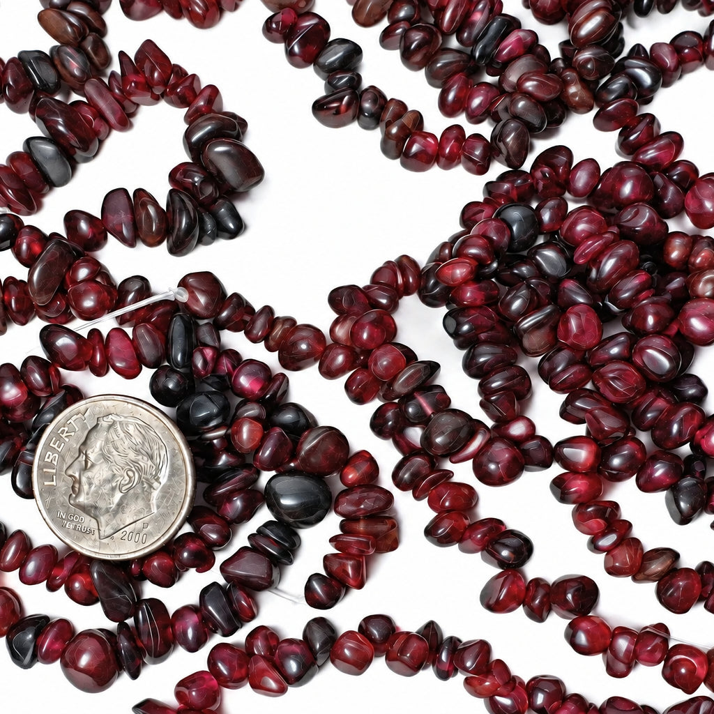 Dark red to black rough gemstone beads draped across a white surface, with a silver coin placed in the center for scale.