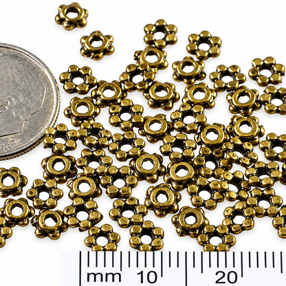 Small metallic flower-shaped findings scattered on a white background, with a coin and a millimeter ruler included for scale.