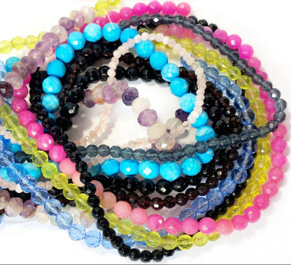 A close-up of multiple beaded bracelets or necklaces arranged in a pile on a white background. The strands feature faceted round beads in bright blue, pink, lime green, black, purple, gray, and translucent light blue and white.