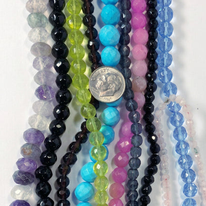 Multiple strands of faceted gemstone beads in various colors, including black, blue, pink, green, purple, and clear, arranged vertically next to a silver coin for scale on a white background.