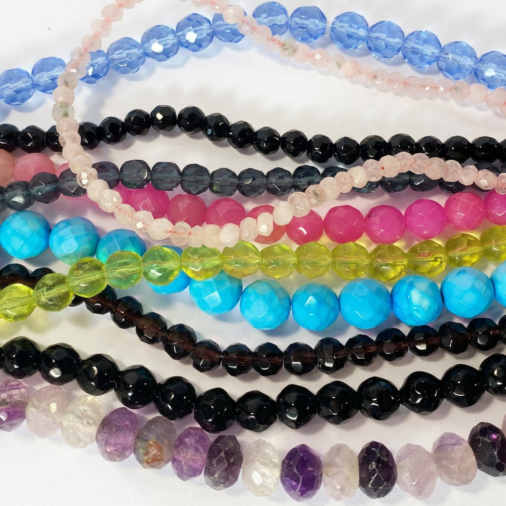 A close-up of multiple strands of faceted gemstone beads in various colors, including blue, pink, black, turquoise, yellow-green, purple, and clear beads, arranged on a white background.