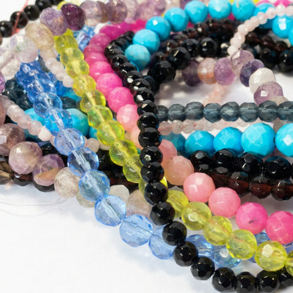 A close-up of multiple strands of faceted round gemstone beads in assorted colors, including turquoise, pink, black, blue, yellow-green, purple, and gray, arranged overlapping on a light surface.