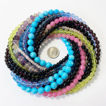 A pile of faceted gemstone beads in multiple colors, including pink, blue, black, yellow-green, purple, and clear, with a coin in the center for scale.